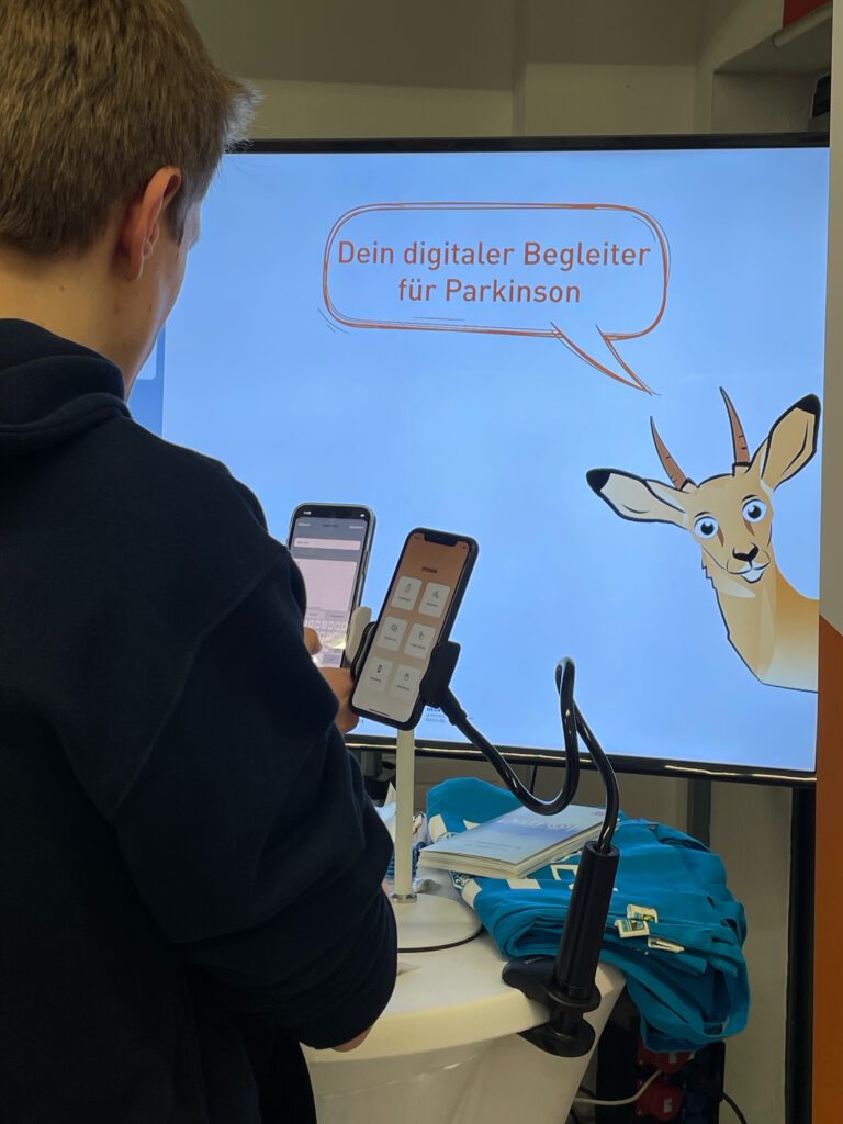 Mann von hinten, der die App Impala an einem Handy testet. Im Hintergrund ein Fernseher mit dem Slogan von Impala